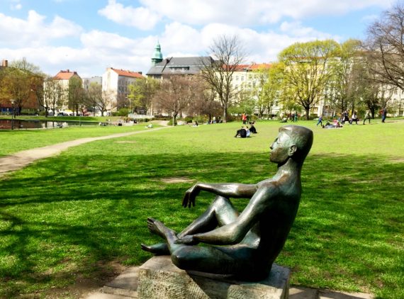 A imagem mostra uma escultura de uma pessoa sentada, meditando, no meio de um gramado de uma praça.