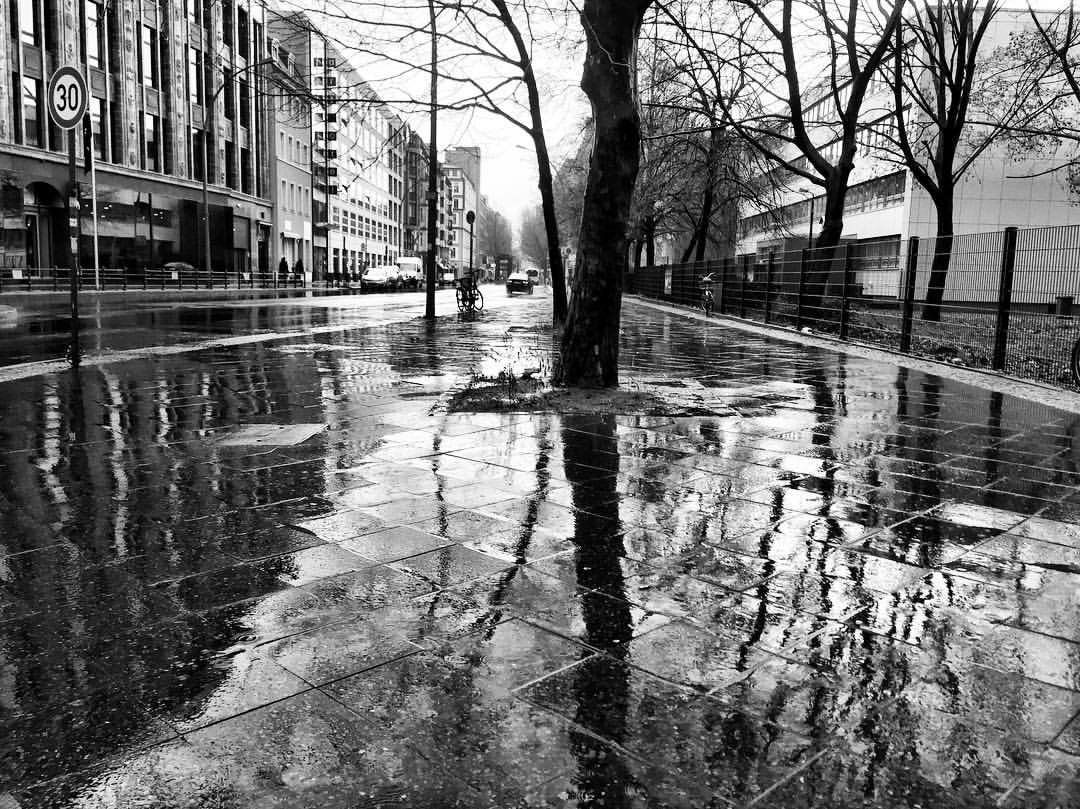 #paracegover A imagem mostra uma foto em preto e branco de uma rua totalmente molhada pela chuva. A calçada reflete os prédios e as árvores sem folhas. — at Märkisches Museum (Berlin U-Bahn).