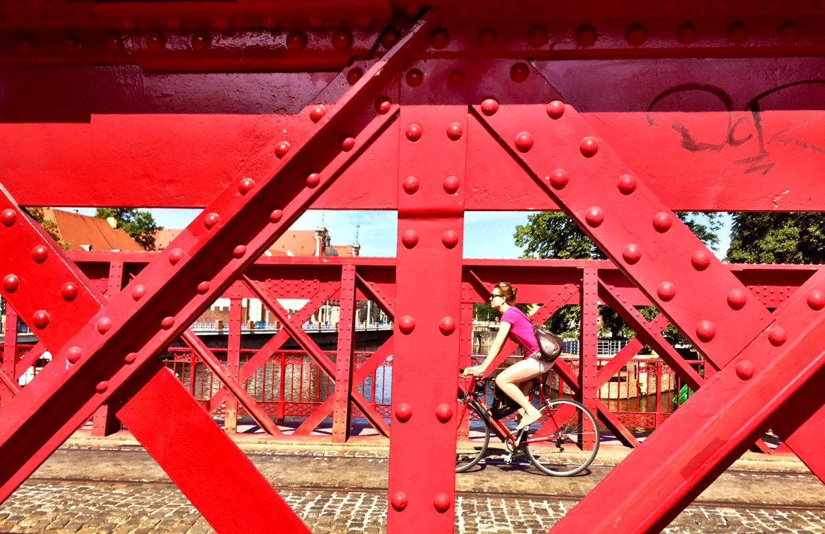 A imagem mostra uma ponte de ferro vermelha enquanto uma ciclista vestida de rosa faz a travessia.