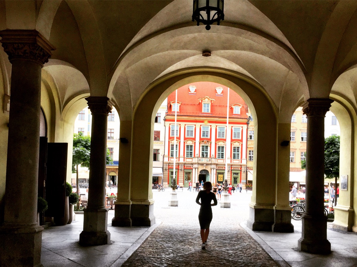 A imagem mostra um portal em forma de arco por onde se vê a silhueta de uma mulher passando. Ao fundo, um prédio vermelho e laranja na praça principal.