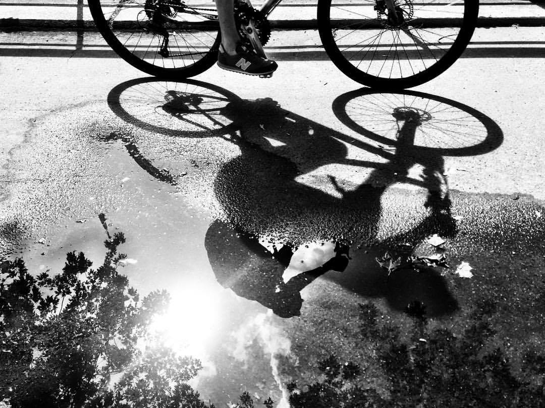 #paracegover Descrição para deficientes visuais: a imagem mostra o reflexo em uma poça d'água da silhueta de uma bicicleta que passa (aparecem apenas as rodas e os pés do ciclista). A foto está em preto e branco para melhorar o contraste. — at Märkisches Ufer.