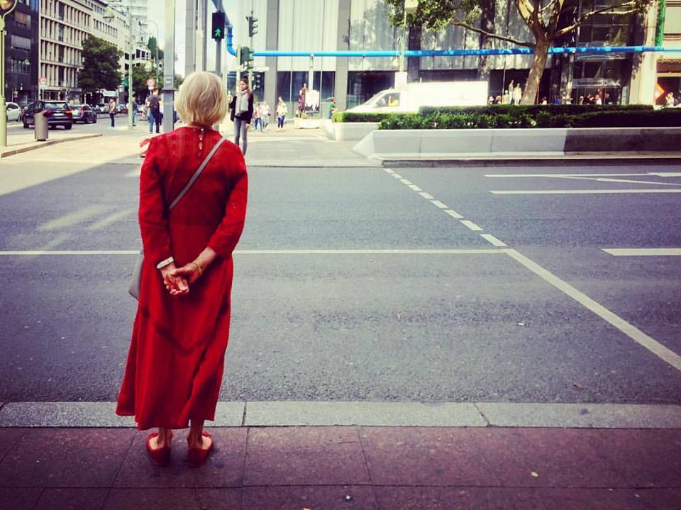 #paracegover Descrição para deficientes visuais: a imagem mostra uma mulher de cabelos brancos vestindo um longo vermelho esperando para atravessar uma avenida larga. Ela está de costas, comas mãos para trás. A rua está praticamente vazia, exceto por um furgão branco que passa do outro lado. — at Kurfürstendamm (Berlin U-Bahn).