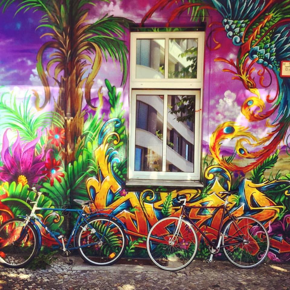 #paracegover Descrição para deficientes visuais: a imagem mostra duas bicicletas estacionadas sob uma janela instalada num prédio cujas paredes da fachada estão grafitadas de maneira espetacular. Cores quentes e intensas, parece que o desenho está vivo e fervendo! — at All Style Tattoo Berlin.