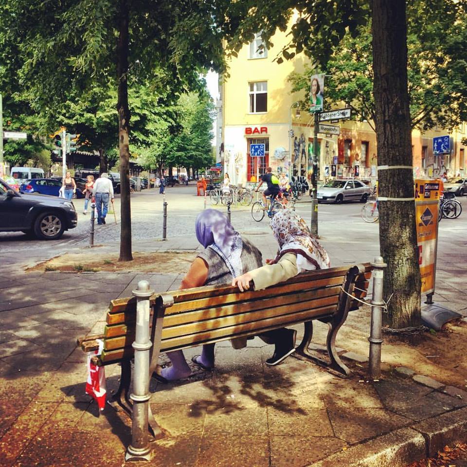 #paracegover Descrição para deficientes visuais: a imagem mostra duas mulheres muçulmanas sentadas num banco de praça observando o movimento. Elas estão de costas embaixo de uma árvore. Pessoas passam caminhando e de bicicleta. — at Estate Coffee.