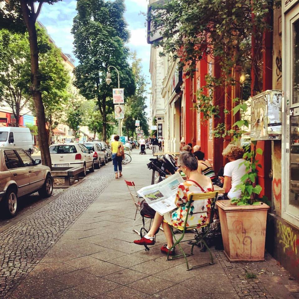 #paracegover Descrição para deficientes visuais: a imagem mostra pessoas num café em mesas na calçada lendo jornal. A rua é arborizada e as paredes do café são laranja escuro. — at Kaffeesatz.