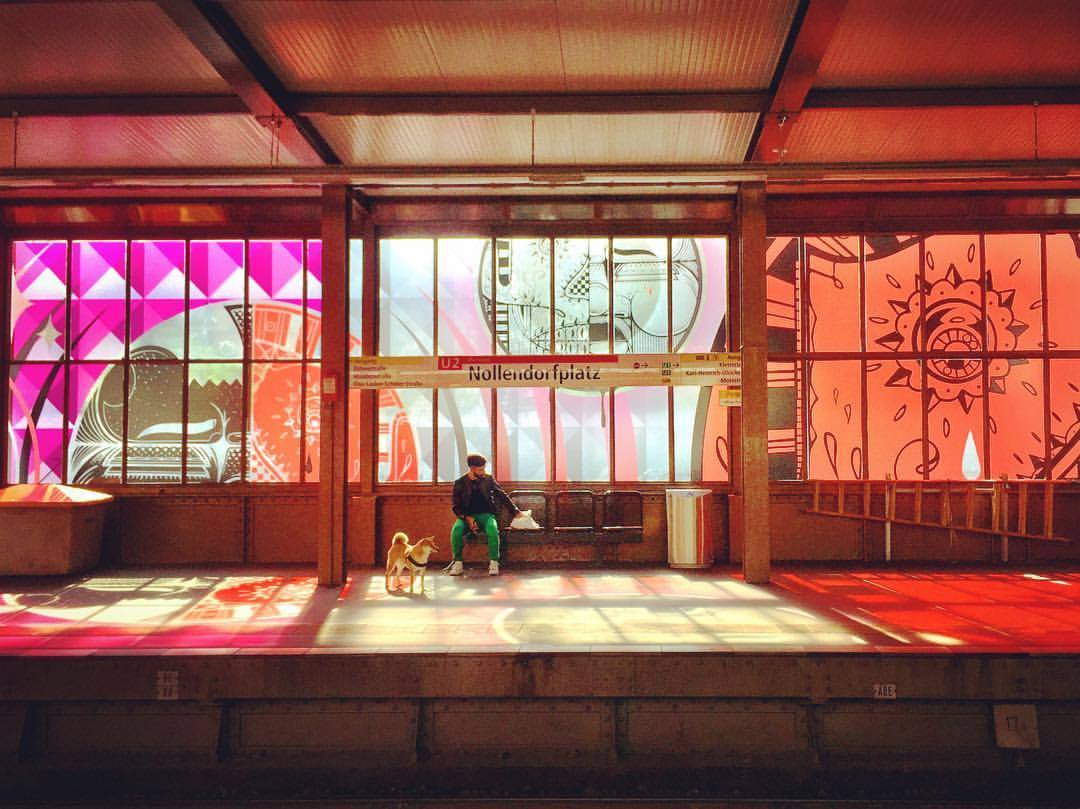 #paracegover Descrição para deficientes visuais: a imagem mostra a estação de metrô Nollendorfplatz cujos vidros foram todos decorados com adesivos transparentes decorados, como uma extensão do projeto do museu de street art, Urban Nation. A luz do sol reflete os desenhos nas cores rosa e laranja. Um homem espera o trem com seu cachorro. — at U-Bahn-Nollendorfplatz.