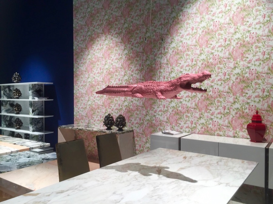 A imagem mostra uma mesa em uma sala com papel de parede estampado com um jacaré rosa usado como luminária.