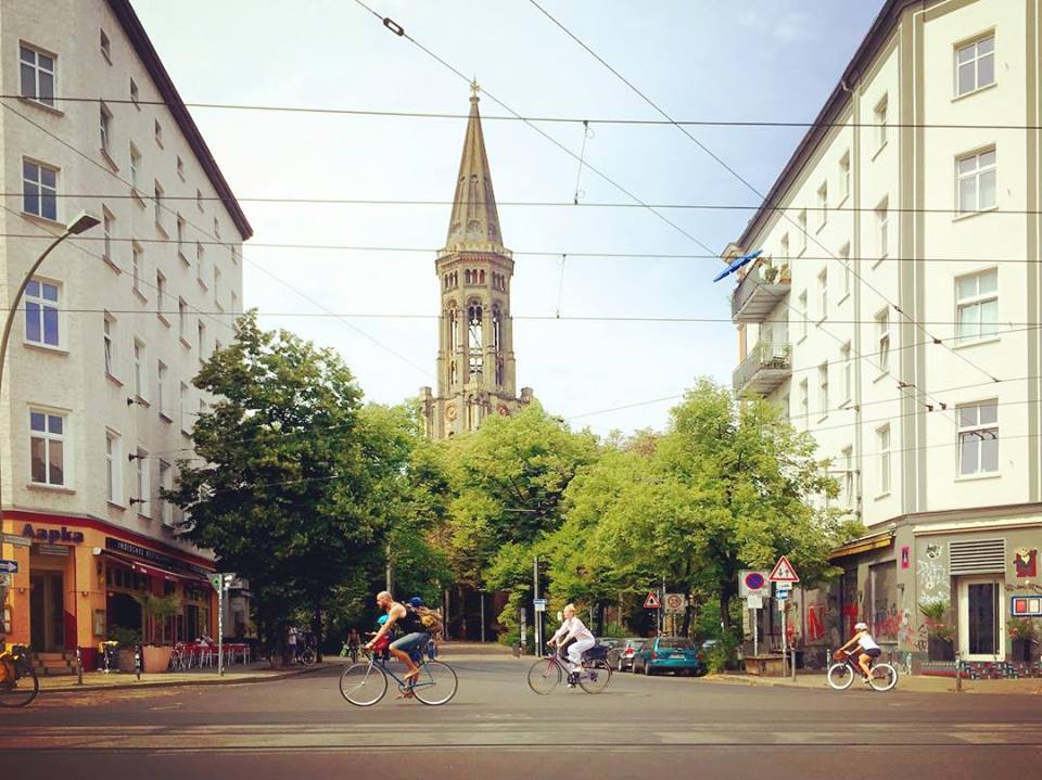 #paracegover Descrição para deficientes visuais: a imagem mostra um cruzamento com uma praça e uma igreja ao fundo. Na rua larga recortada com os trilhos do tram, passam bicicletas apressadas. — at Zionskirchstraße.