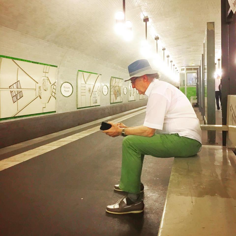 #paracegover Descrição para deficientes visuais: a imagem mostra um senhor cheio de estilo sentado no banco de madeira da estação de metrô, futucando seu celular. Ele usa camisa branca, calça verde-bandeira, mocassins bicolores e um chapéu de tecido. — at Märkisches Museum (Berlin U-Bahn).
