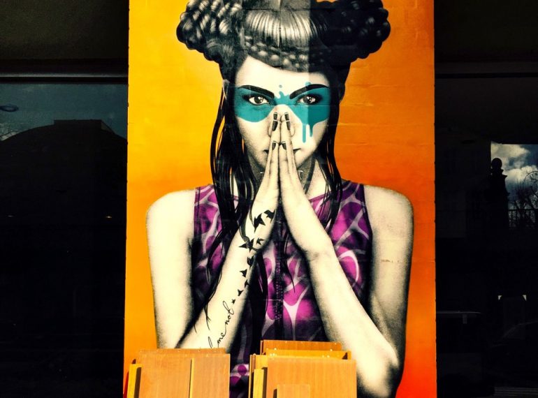 A imagem mostra a foto de um grafite feito pelo artista Finnbarr Dac, que mostra uma mulher com uma palma da mão de encontro à outra, em posição medidativa. Ela usa penteado e maquiagem bem exóticos, mas belíssimos.