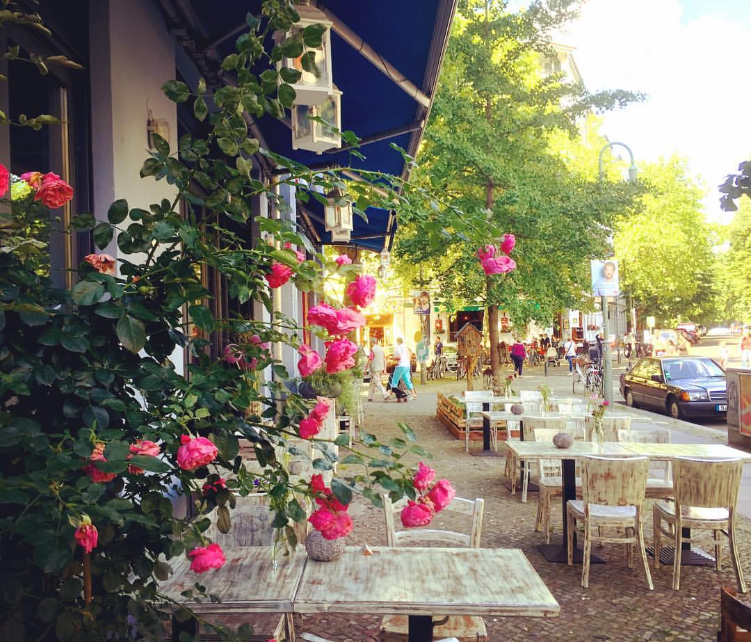 5. Ruas cheias de rosas, lindas, coloridas e perfumadas. Como não amar? #paracegover A imagem mostra as mesas brancas de um café numa calçada larga. Ao fundo, árvores frondosas. O quadro é emoldurado por mini rosas cor-de-rosa. O dia está lindo (Knaackstraße).