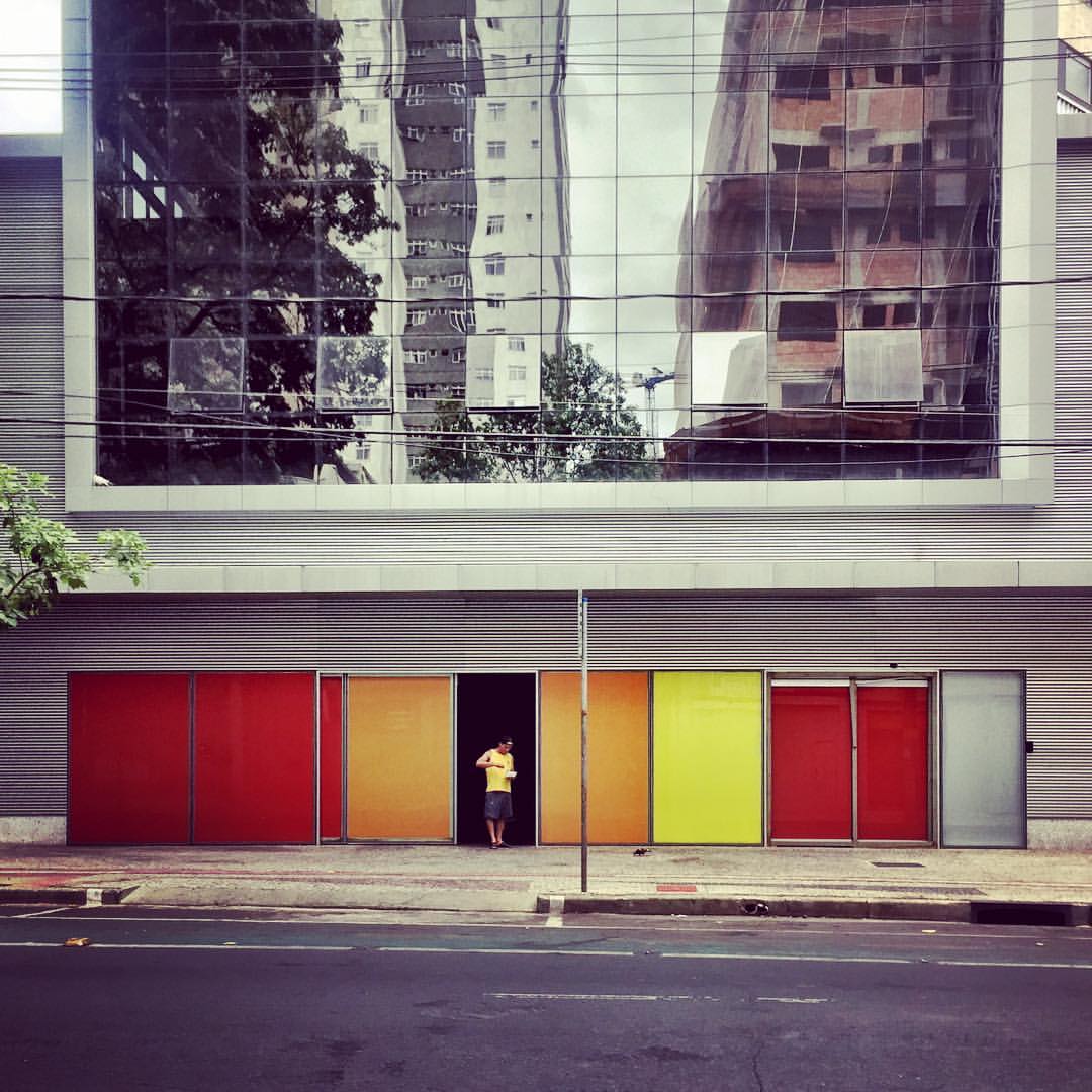 A imagem mostra a fachada de um prédio com superfícies nas cores vermelho, laranja e amarelo. Uma pessoa de camiseta amarela está na porta. A parte superior é de vidro espelhado. — at Circus Rock Bar.