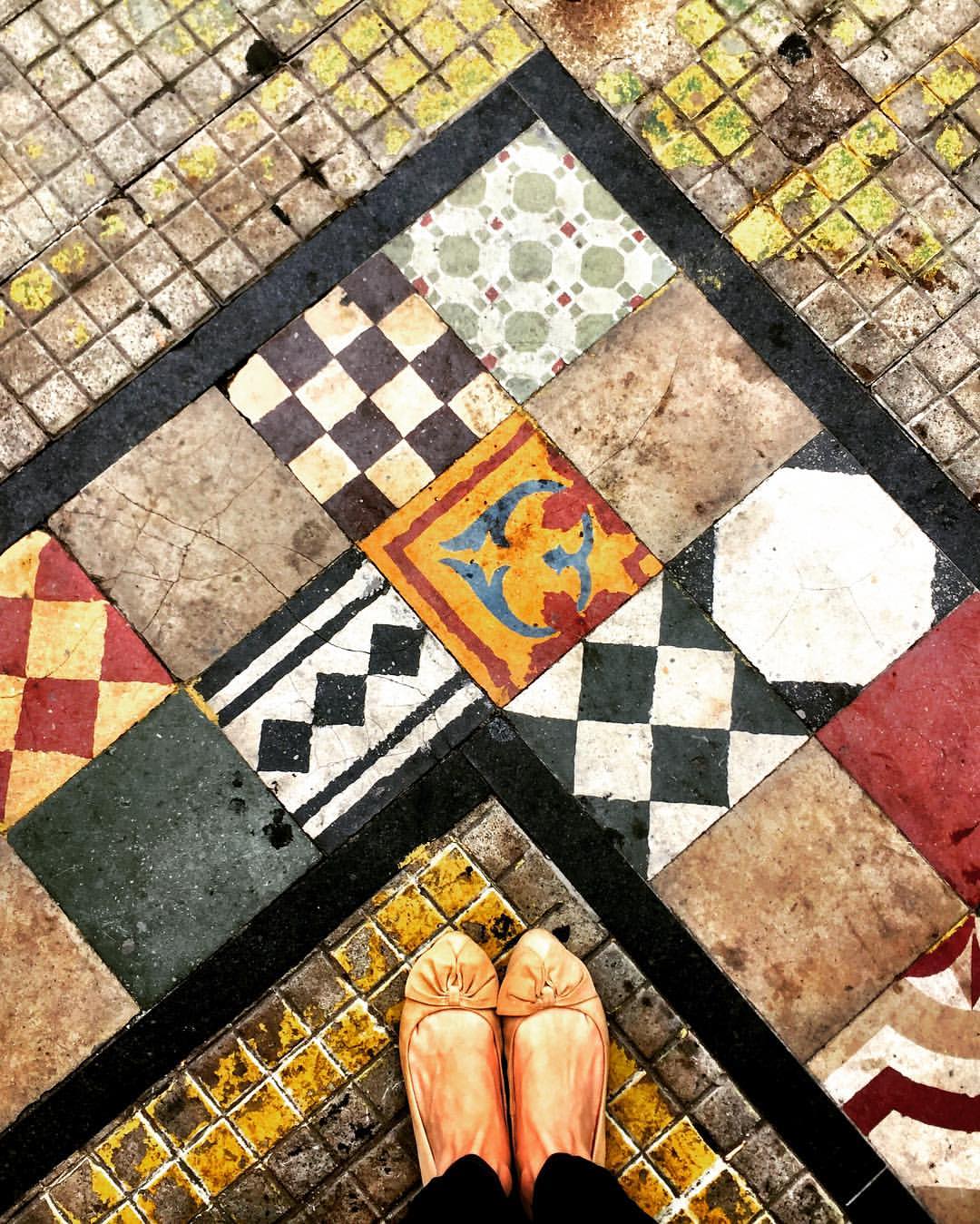 #paracegover A imagem mostra uma calçada decorada com azulejos hidráulicos coloridos, formando um ângulo reto. Meus pés aparecem no canto inferior, na base do triângulo. — at Boi lourdes.