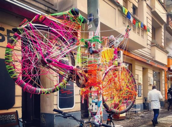 #paracegover A imagem mostra uma bicicleta pendurada num poste, toda coberta por linhas de cores diversas, fazendo as vezes de uma escultura, totalmente integrada ao ambiente. Uma das sacadas do prédio que aparece ao fundo é enfeitada com bandeirinhas coloridas. O povo desse bairro (Friedrischshain) gosta de cenários coloridos (eu também). — at Gabriel-Max-Straße.