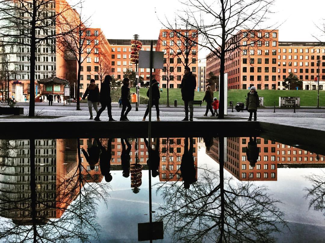 #paracegover A imagem mostra um grupo de pessoas à beira de um espelho d' água. Os prédios que aparecem ao fundo, assim como as árvores sem folhas são refletidas também. — at Potsdamer Platz.