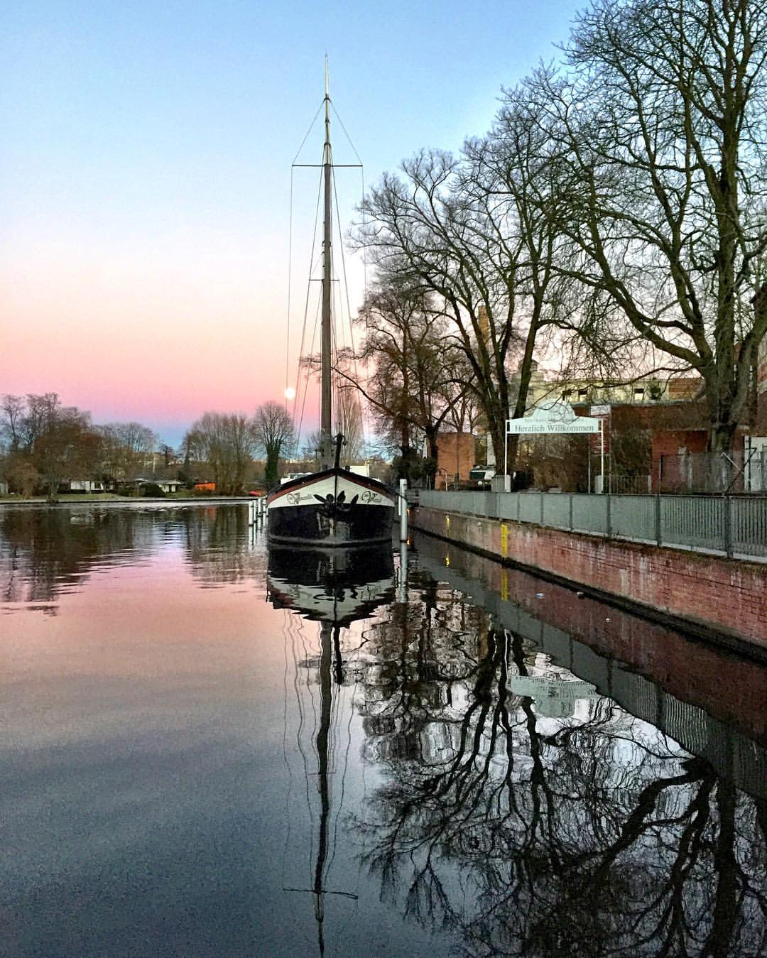  #paracegover A imagem mostra um barco a vela refletido na água na hora do crepúsculo. O céu vai do azul intenso, passando pelo laranja e chegando em vários tons de rosa. Se olhar com atenção, é possível perceber a lua atrás do mastro. — at Berlin Köpenick Spree Ufer.