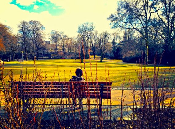 A imagem mostra uma pessoa sentada num banco. Ela é vista de costas, de frente para um amplo gramado num parque.