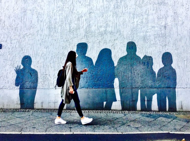 #paracegover A imagem mostra uma moça andando numa calçada. No muro atrás dela há uma série de silheutas de pessoas pintadas. Parecem sombras.