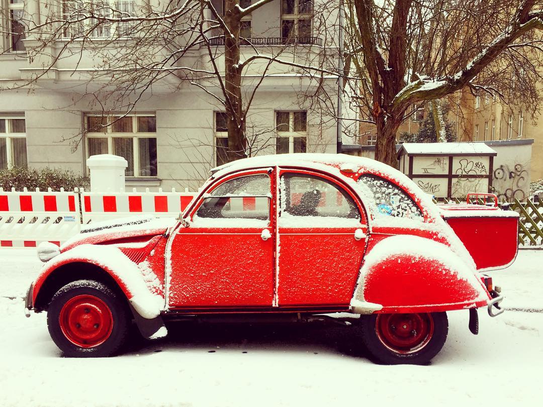 Descrição para deficientes visuais: a imagem mostra um Citröen antigo estacionado na rua. O vermelho intenso do carro da mesma cor dos detalhes da cerca de proteção atrás contrasta com a neve que cobre tudo. — at Volkspark Schöneberg