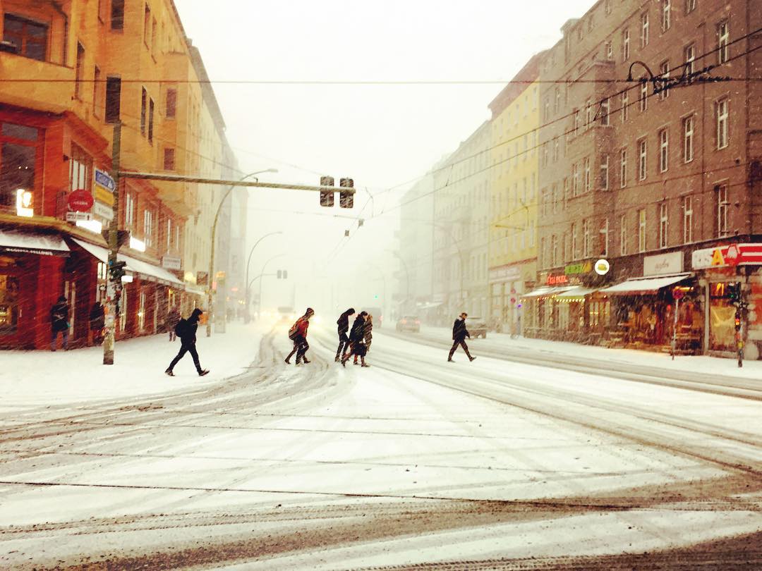 Descrição para deficientes visuais: a imagem mostra pessoas atravessando a rua coberta de neve. Em contraste com o frio intenso, os prédios nas laterais são pintados com cores quentes, como vermelho, laranja e amarelo. — at Eberswalder Straße.