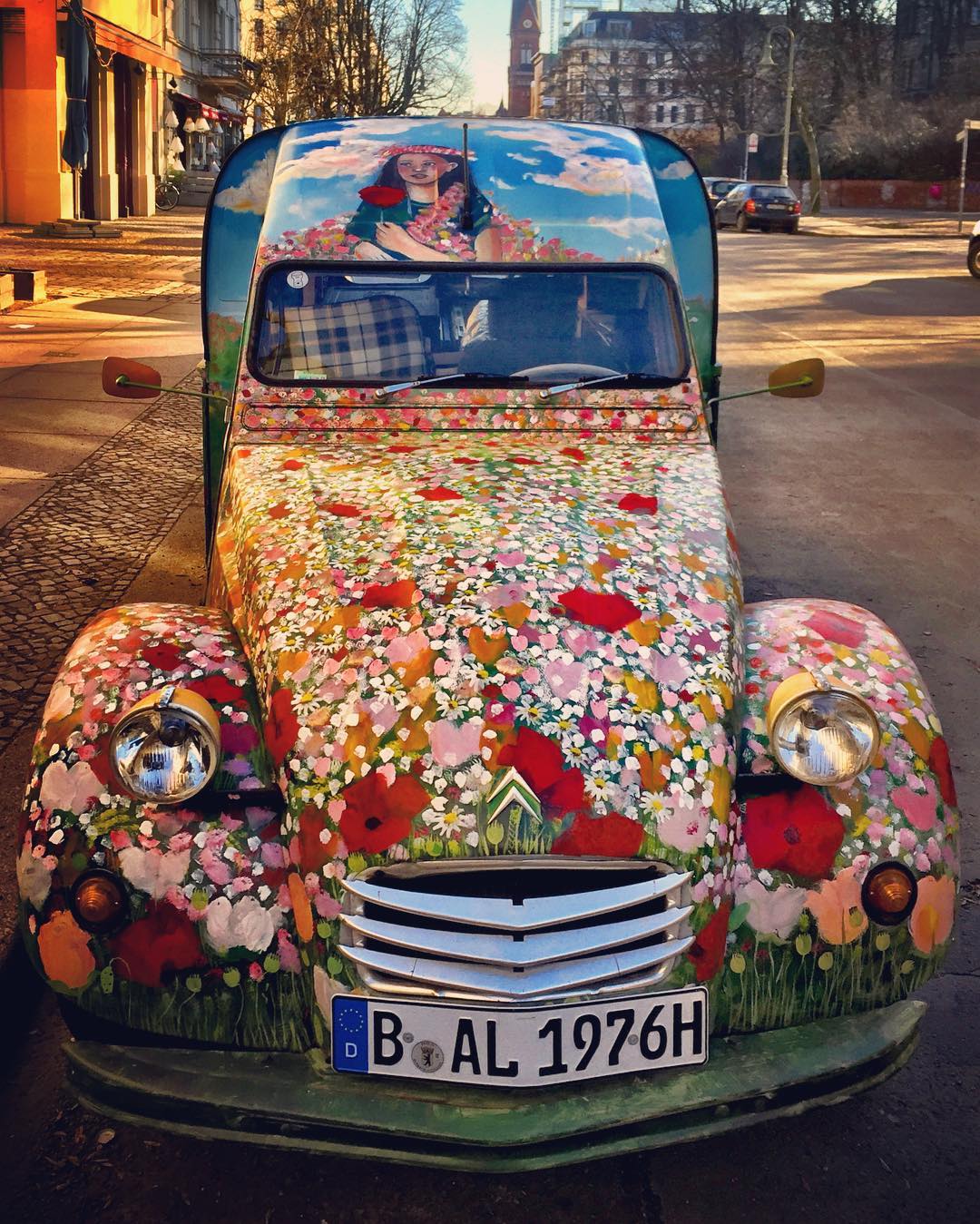 Descrição para deficientes visuais: a imagem mostra um carro antigo visto de frente (acho que é um Citröen) todo estampado com flores do campo. Na capota, o desenho de uma moça de cabelos pretos segurando uma rosa vermelha. O veículo é do restaurante francês Paulette, que fica no local onde ele está estacionado. Primavera ambulante � — at Knaackstraße.