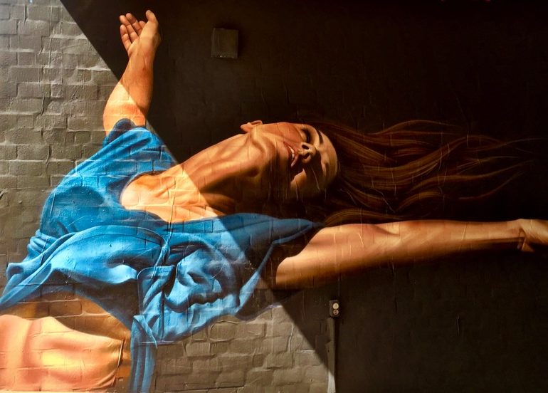 A imagem mostra um grafite hiperrealista de uma mulher se alongando com os braços abertos e uma blusa azul.