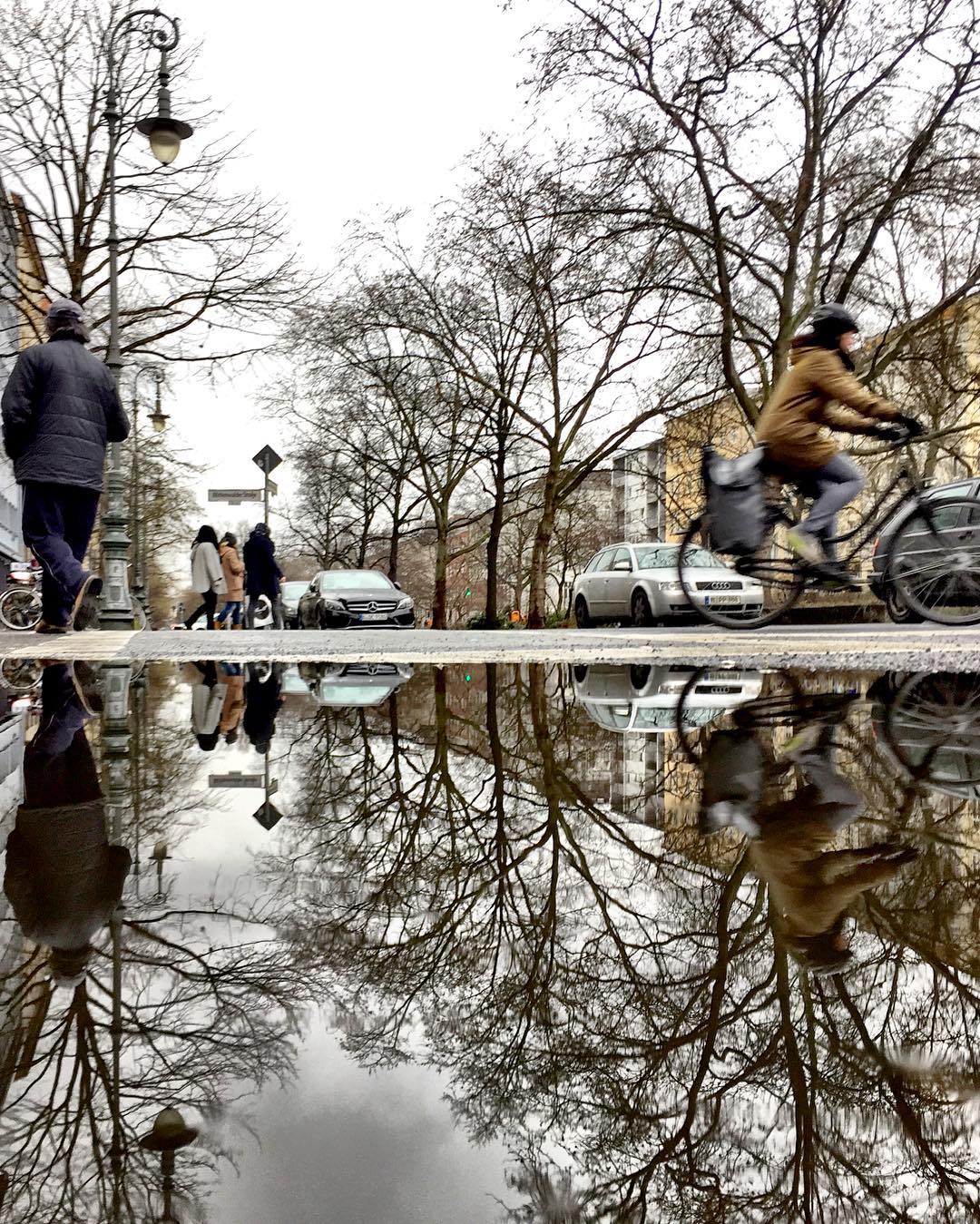 #paracegover Descrição para deficientes visuais: a imagem mostra uma rua onde passam pessoas, carros e ciclistas. Tudo, incluindo o poste de iluminação, o céu nublado e as árvores sem folhas, está refletido na poça d'água em primeiro plano. — at Gneisenaustraße.