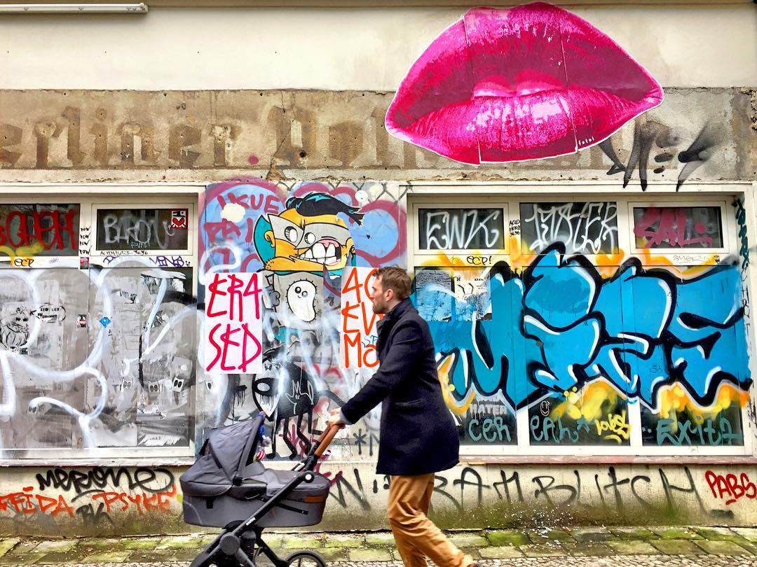 #paracegover Descrição para deficientes visuais: a imagem mostra um homem caminhando com um carrinho de bebê. Ao fundo, uma parede toda grafitada com um grande beijo pink em destaque. — at Gabriel-Max-Straße.