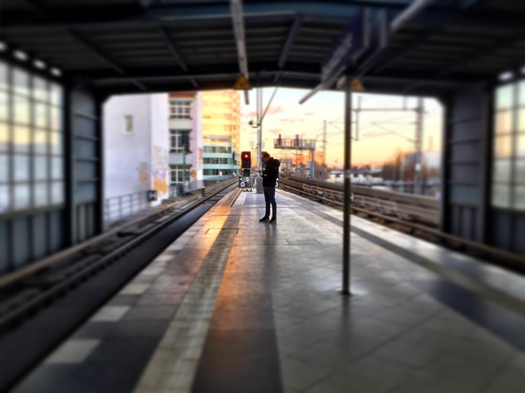 #paracegover Descrição para deficientes visuais: a imagem mostra uma estação de trem vista por dentro. Há uma pessoa na plataforma vazia, consultando seu celular. A luz do sol faz com que os prédios ao fundo fiquem dourados. — at Berlin Jannowitzbrücke station.