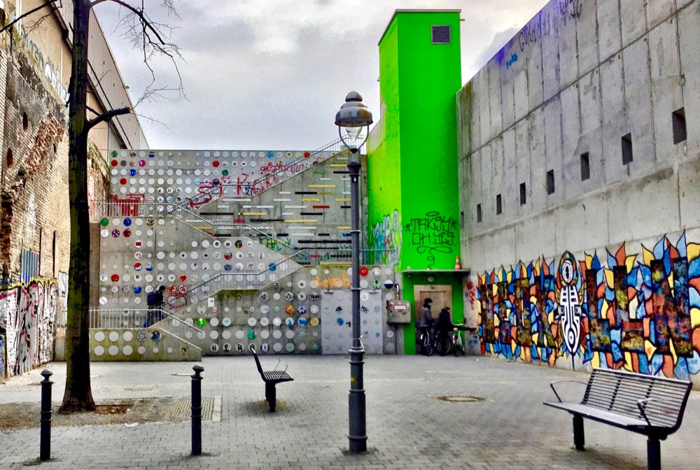 A imagem mostra um beco com uma escadaria de concreto no final, cheia de intervenções artísticas. Do lado direito há um elevador pintado de verde-limão. 