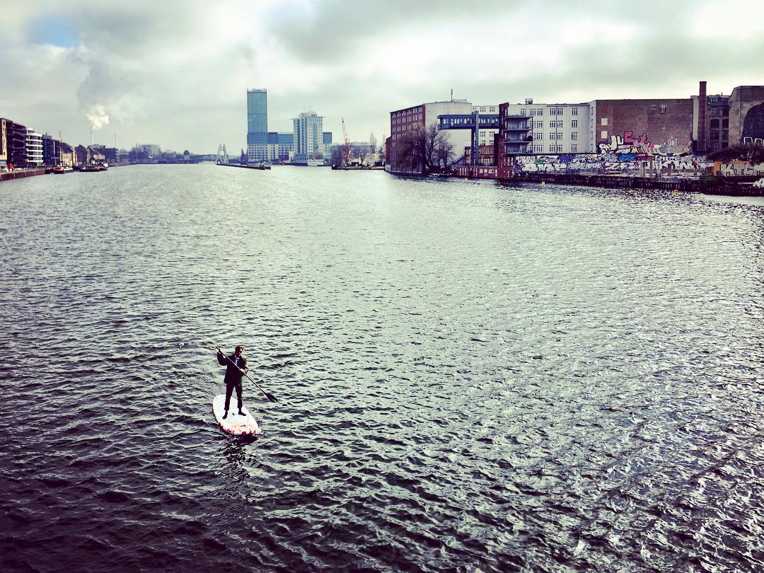 #paracegover Descrição para deficientes visuais: a imagem mostra um homem solitário no meio do rio Spree praticando Paddling (remo em pé sobre uma prancha parecida com a de surf). Ao fundo, prédios e a escultura Molecule Man podem ser vistos. — at Oberbaum Bridge.