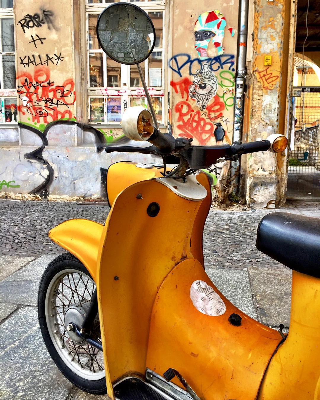 #paracegover Descrição para deficientes visuais: a imagem mostra a parte dianteira de uma vespa amarela estacionada na calçada. Atrás dela, uma parede grafitada com vários desenhos. A foto foi tirada de maneira a parecer que o passarinho pintado na parede está pousado no guidão da Vespa. Parece que ele quer passear; vamos? — at Prater Garten.