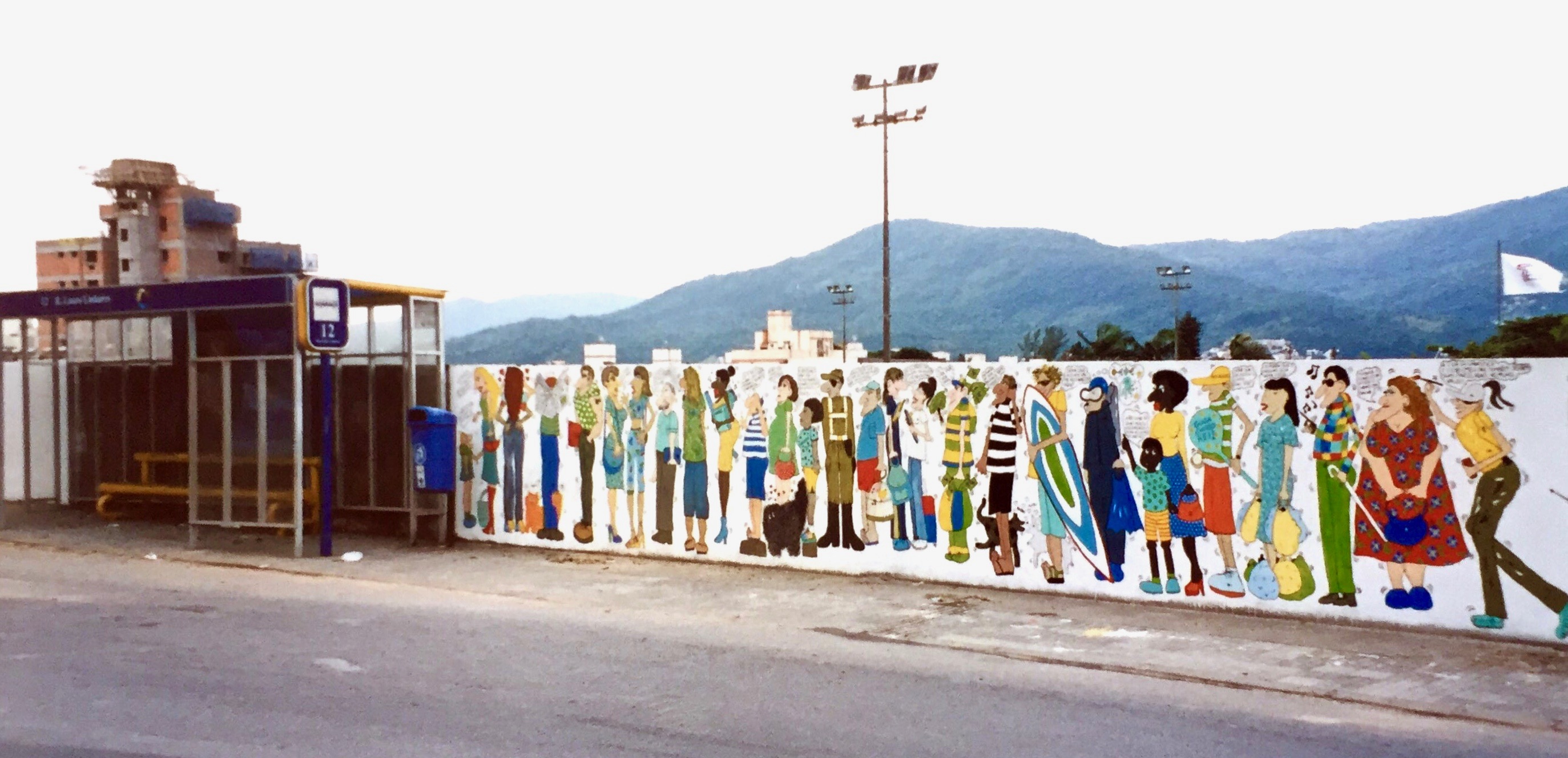 #paracegover A imagem mostra um ponto de ônibus coberto.No muro ao redor dele, personagens desenhados conversando entre si.