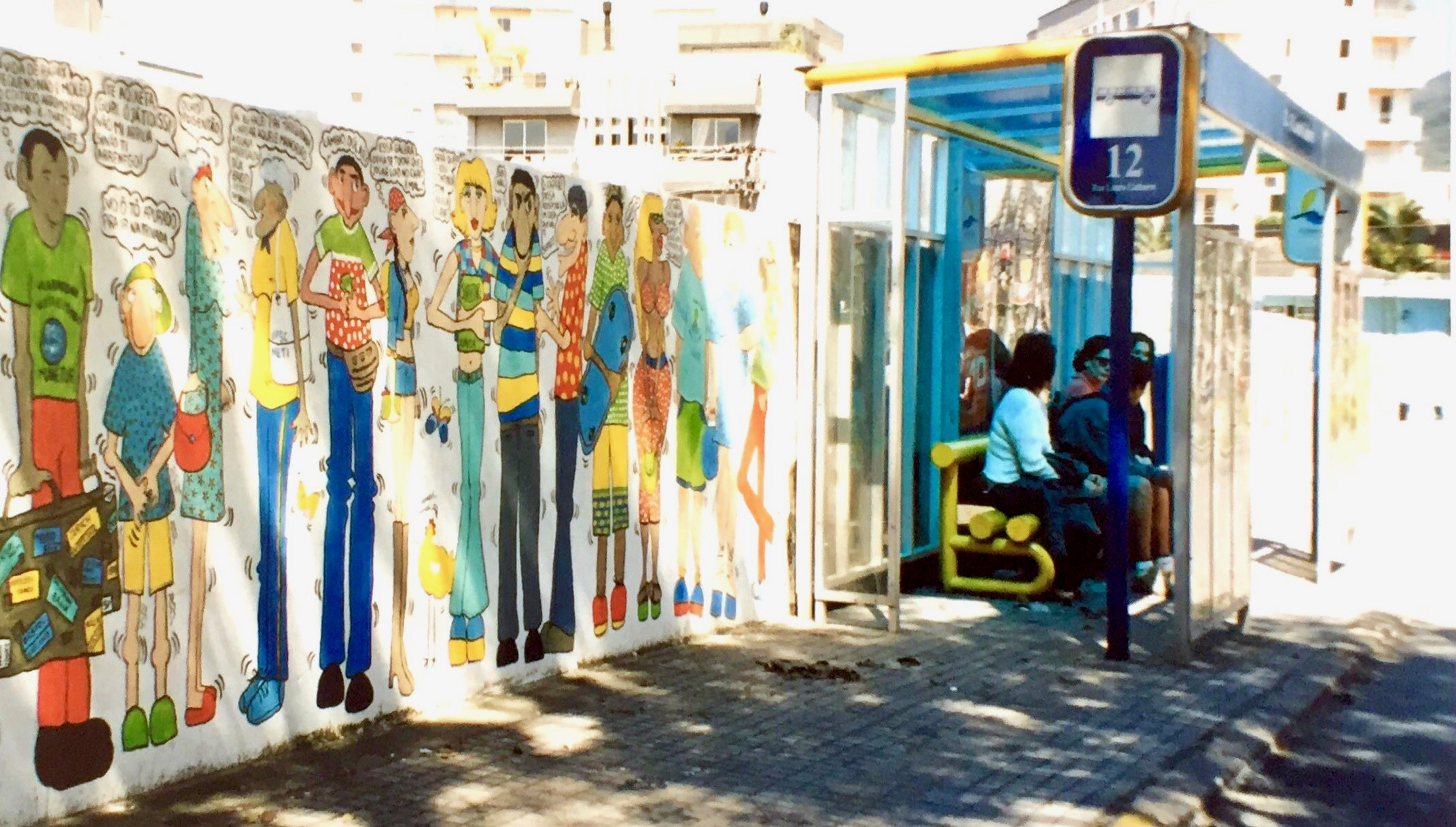 #paracegover A imagem mostra pessoas sentadas no ponto de ônibus ao lado do muro desenhado.