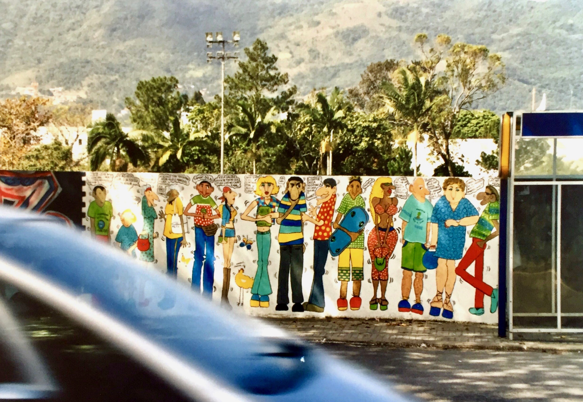 #paracegover A imagem mostra um muro desenhado com pessoas em volta de um ponto de ônibus. A foto foi tirada do outro lado da rua e um carro passou bem na hora.