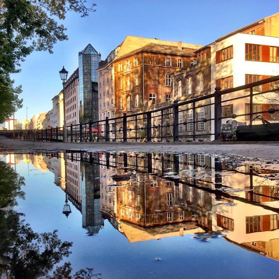 #paracegover Descrição para deficientes visuais: a imagem mostra um conjunto de prédios banhados pela luz dourada do sol, às margens de um dos canais do rio Spree. A cena é refletida numa poça d'água. — at Derag Hotel und Living Grosser Kurfuerst Berlin.