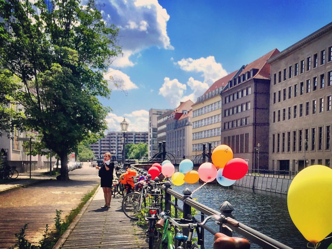 #paracegover Descrição para deficientes visuais: a imagem mostra bicicletas estacionadas ao longo da grade de proteção de um canal do rio Spree. Balões de gás coloridos trazem alegria e cor ao cenário. Uma pessoa está parada na calçada tirando fotos com um iPad (mas de costas para os balões!). O dia está lindo. — at Friedrichsgracht.