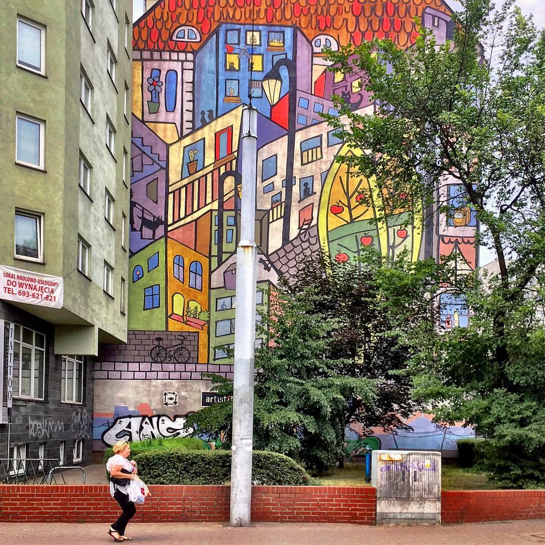  #paracegover Descrição para deficientes visuais: a imagem mostra uma mulher caminhando apressada por uma calçada em frente a um jardim. Ao fundo, uma parede cega decorada com um mural coloridíssimo, retratando um conjunto de prédios em estruturas geométricas.