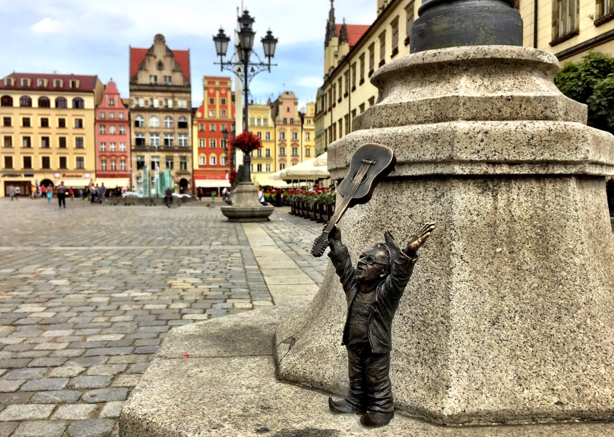 A imagem mostra uma pequena escultura em bronze de um homenzinho mostrando um violão com as mãos para cima. Ao fundo, o casario da praça principal.