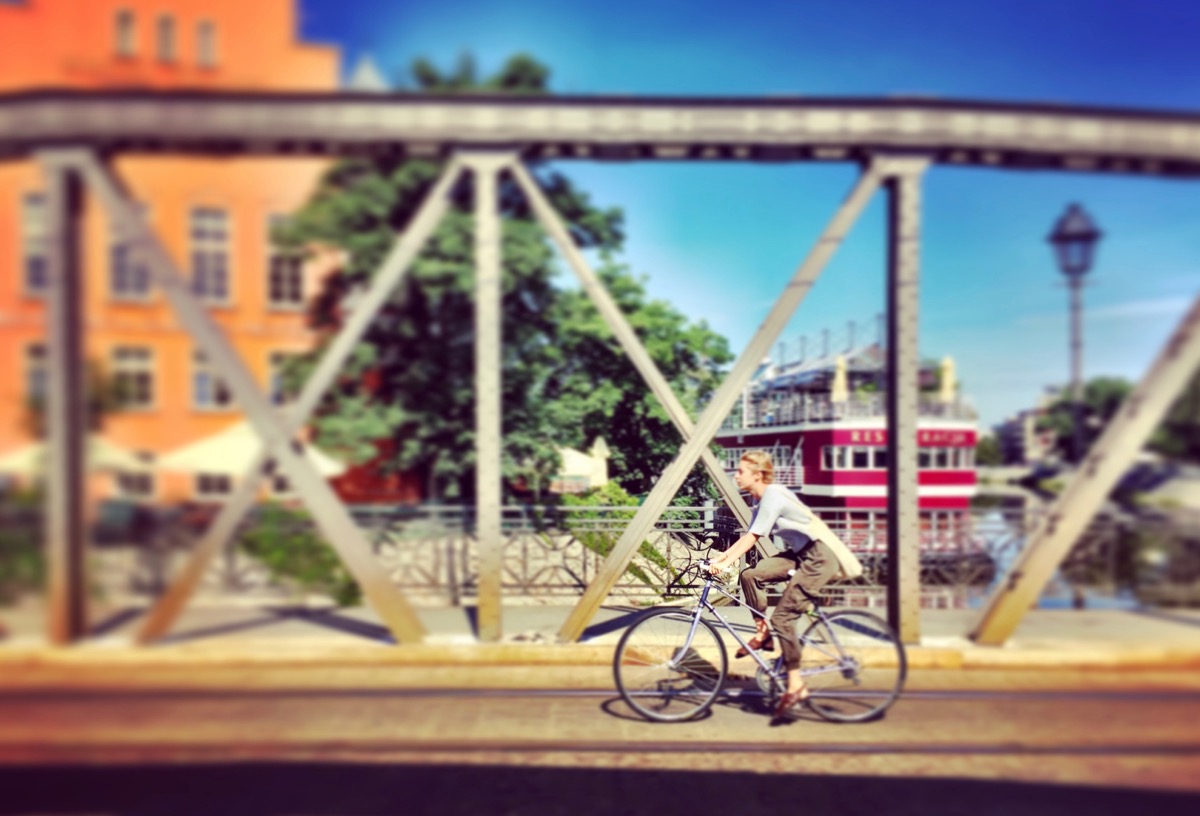 A imagem mostra uma mulher passando de bicicleta por uma ponte de ferro.