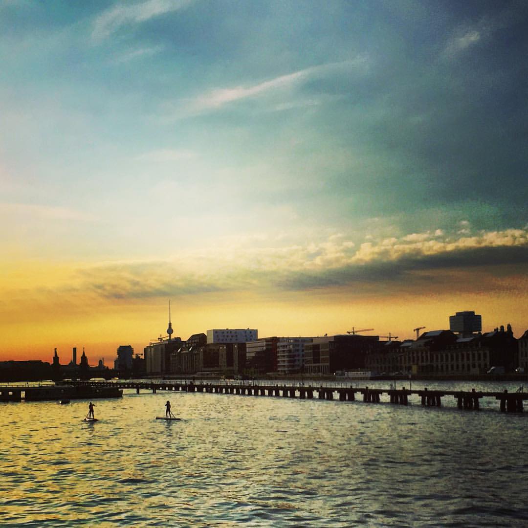 #paracegover Descrição para deficientes visuais: a imagem mostra um por-do-sol espetacular visto de uma margem do rio Spree. Ao fundo, na skyline da cidade, é possível ver a torre de tv. Duas pessoas fazem stand up paddle. — at Molecule Man (sculpture).