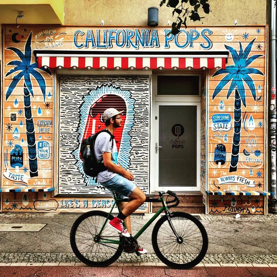 #paracegover Descrição para deficientes visuais: a imagem mostra um rapaz passando de bicicleta na ciclovia bem em frente a uma sorveteria, que está fechada no momento. A fachada é toda decorada com desenhos e dizeres que remetem ao verão. Há um coqueiro azul desenhado em cada lado da fachada. — at California Pops - Prenzlauer Berg.