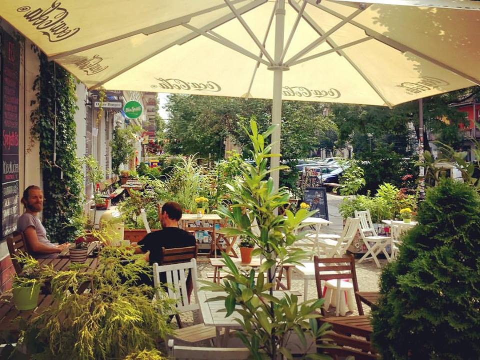 #paracegover Descrição para deficientes visuais: a imagem mostra as mesas de um café na calçada larga e arborizada. Um ombrelone protege os frequentadores do sol. É a paz materializada em paisagem urbana. — at Café Entweder Oder.