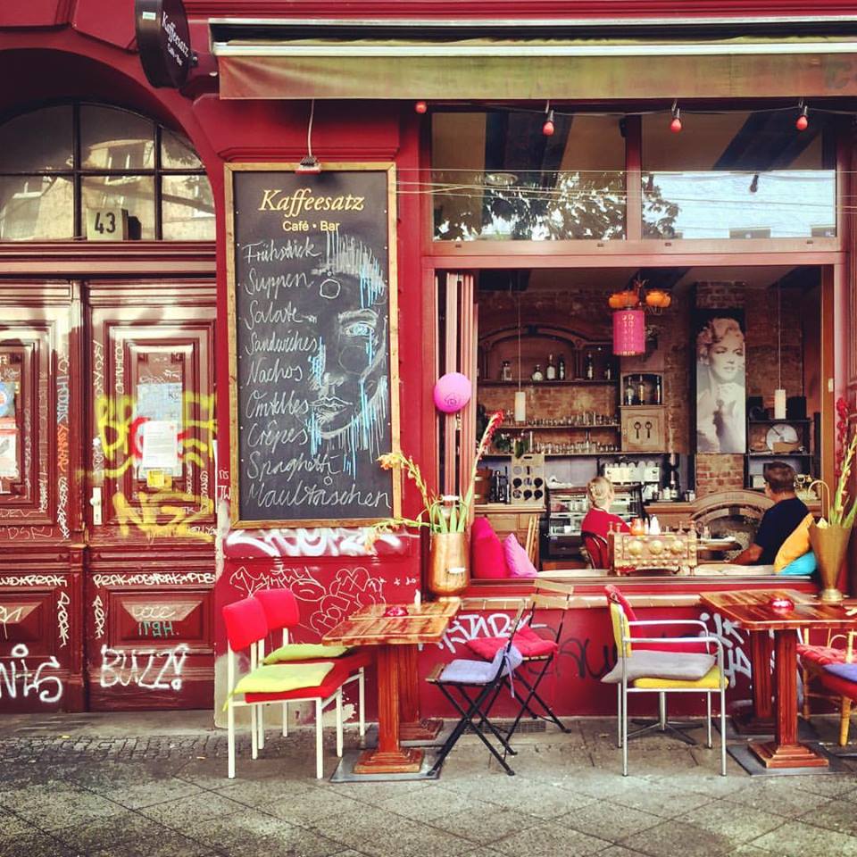 #paracegover Descrição para deficientes visuais: a imagem mostra a fachada de um belíssimo café; as paredes são de um vermelho intenso e as almofadas das cadeiras na calçada e são pink, amarelo, verde-limão e lilás. Os vasos são cor de cobre. Pela janela pode-se ver dois clientes sentados do lado de dentro: um homem veste camiseta preta e uma mulher está de blusa vermelha. — at Kaffeesatz.