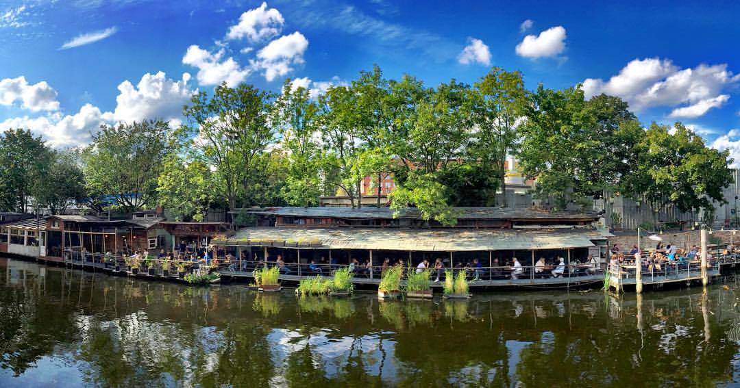 #paracegover Descrição para deficientes visuais: a imagem panorâmica mostra o restaurante FreiSchwimmer visto da margem oposta de um dos canais do rio Spree. A vegetação é densa, próxima do Treptower Park. A cidade mais verde que conheci e que mora no meu coração. Berlim, sua linda! ❤️ — at Freischwimmer.