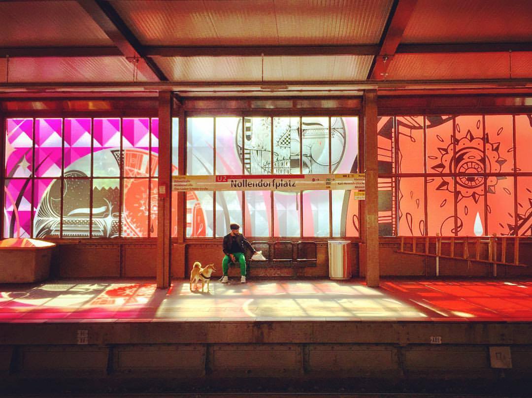 #paracegover Descrição para deficientes visuais: a imagem mostra a estação de metrô Nollendorfplatz cujos vidros foram todos decorados com adesivos transparentes decorados, como uma extensão do projeto do museu de street art, Urban Nation. A luz do sol reflete os desenhos nas cores rosa e laranja. Um homem espera o trem com seu cachorro. — at U-Bahn-Nollendorfplatz.
