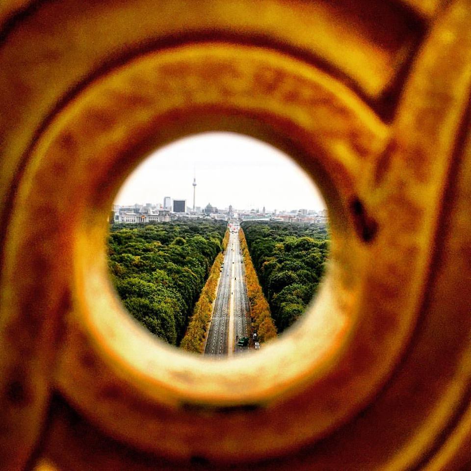 #paracegover Descrição para deficientes visuais: a imagem mostra a skyline de Berlim vista a partir de um dos furos da cerca de proteção no alto da coluna da Vitória. A Avenida 17 de junho aparece ladeada por árvores que já estão amarelas, em contraste com o enorme parque, cuja maioria das árvores ainda está bem verde. Ao fundo, o Portão de Brandemburgo e a Torre de TV. — at Berlin Victory Column.