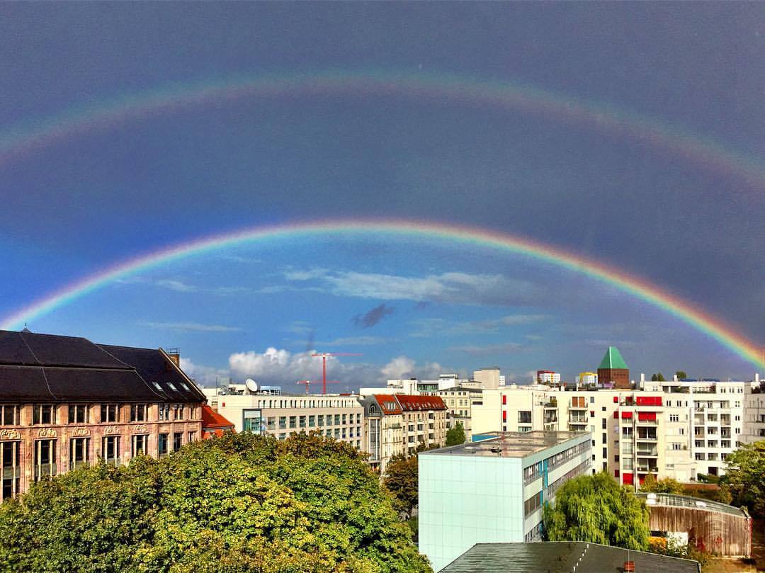 #paracegover Descrição para deficientes visuais: a imagem mostra um arco-íris duplo. Na parte de baixo, o céu está azul; na parte de cima, bem nublado. Também aparecem prédios e árvores. — at Berlin Mitte.