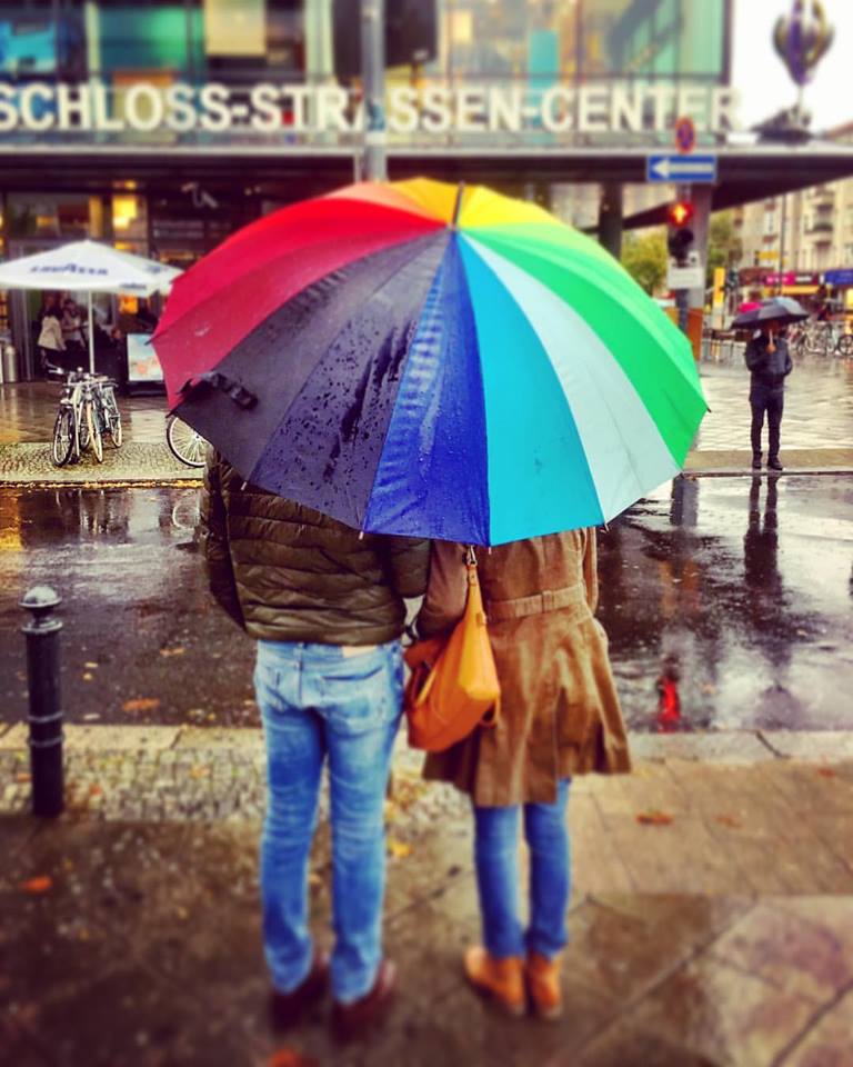 #paracegover Descrição para deficientes visuais: a imagem mosyra um casal de costas, esperando o sinal abrir para atravessar a rua. Eles estão protegidos por um enorme guarda-chuva com gomos coloridos. Chove. — at Schloßstraßen Center.