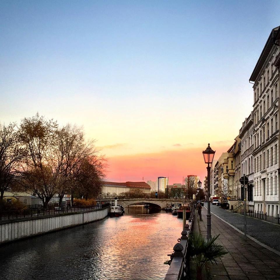  #paracegover Descrição para deficientes visuais: a imagem mostra um canal do rio Spree com uma ponte. Nas laterais, edifícios antigo e uma luminária charmosa. O céu está parte azul e parte rosa pêssego.